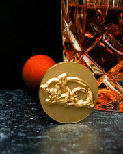 A close-up image of a golden ice stamp with an embossed design resting against a cut-glass tumbler filled with an amber-colored liquid. 