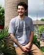 A model sitting against a palm tree with his hands together, wearing a gray T-shirt with a circular emblem in the center with the text "T-REX" at the top, "ORLANDO" at the bottom, and "NEVER SKIP ARM DAY, EST 08" in the center in a black ribbon, with a design of a T-rex lifting weights above it. 