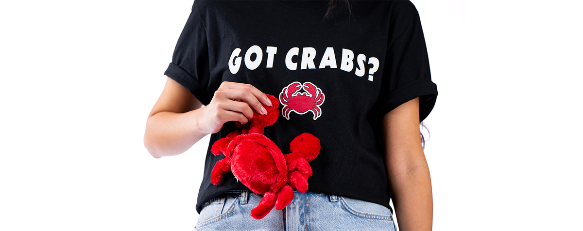 The image features a person holding a red plush crab toy and wearing a black T-shirt with the humorous text “GOT CRABS?” in bold white letters, accompanied by a stylized image of a red crab. The person is also wearing light blue denim jeans, and the background is plain white, highlighting the person and their playful attire.