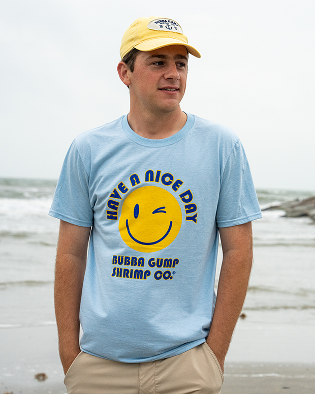 A young man stands on a beach with his hands in khaki bottoms, wearing a light blue shirt with a smiley face and the text "Have a Nice Day, Bubba Gump Shrimp Co." written in navy surrounding it. He is also wearing a yellow baseball cap with a white circular patch, navy lettering and an anchor design on it.
