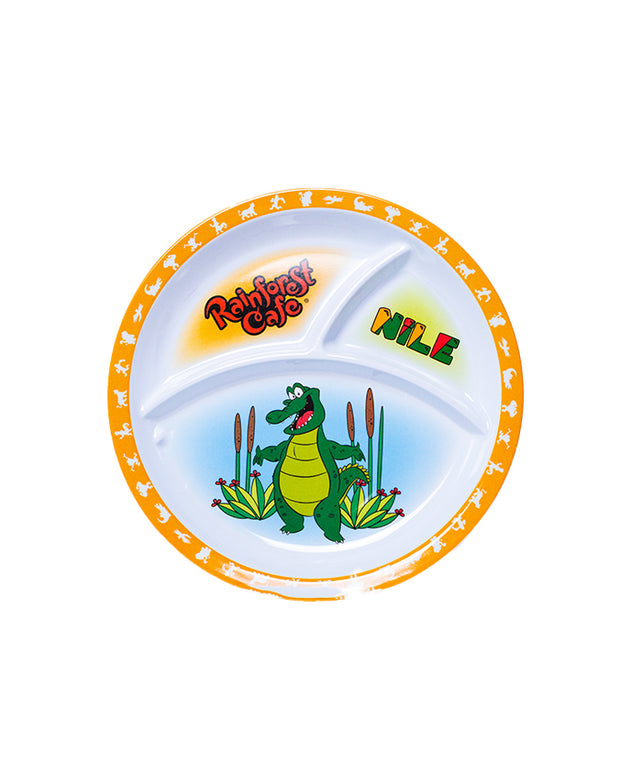 A colorful children’s divided plate featuring a cartoon illustration. The main section of the plate shows a green crocodile character standing happily with a pile of green plants behind it. Above the crocodile, the text ‘Rainforest Cafe’ is written in stylized, playful letters with leaf motifs. The smaller section of the plate has the word ‘NILE’ written in bold, colorful block letters against a white background.