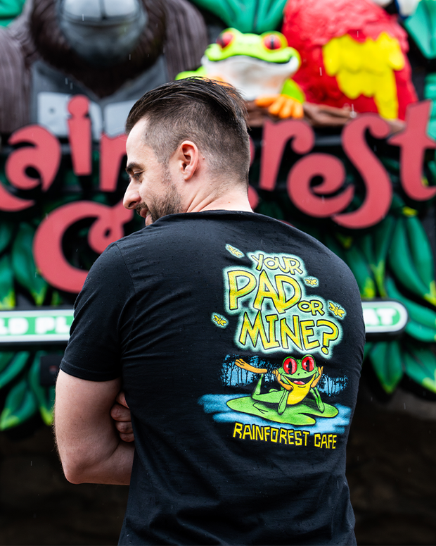 A model with their back to the camera, wearing a black t-shirt that has a colorful design on the back. The design features a cartoon frog sitting on a lily pad with the text "YOUR PAD OR MINE?" above it and "RAINFOREST CAFE" below it in yellow letters. In the background, there is a sign for a Rainforest Cafe Restaurant.
