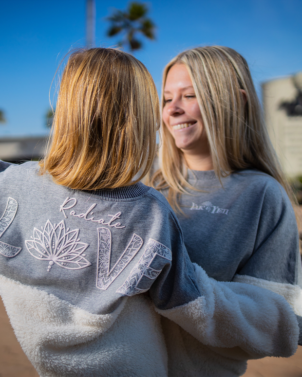 The image shows two individuals standing side by side with one facing to the camerea and the others back is to the camera. The person on the left has shoulder-length hair and is wearing a grey sweatshirt with the word “Radiate” in stylized text and the word "Love" with a lotus flower graphic as the "O" . The person on the right has long hair and is wearing a grey sweatshirt under a white fluffy jacket. 
