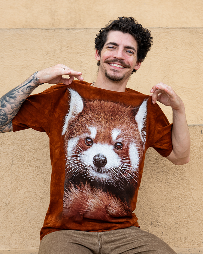 A close image of a model sitting against a sandstone background with a brownish red T-shirt with a realistic red panda face on it with its tail curled under it. 