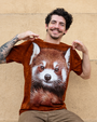 A close image of a model sitting against a sandstone background with a brownish red T-shirt with a realistic red panda face on it with its tail curled under it. 