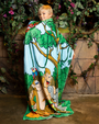 The backside view of a little boy in a jungle setting, wrapped in a colorful blanket featuring cartoon animals in a jungle scene, surrounded by green plants and vines.