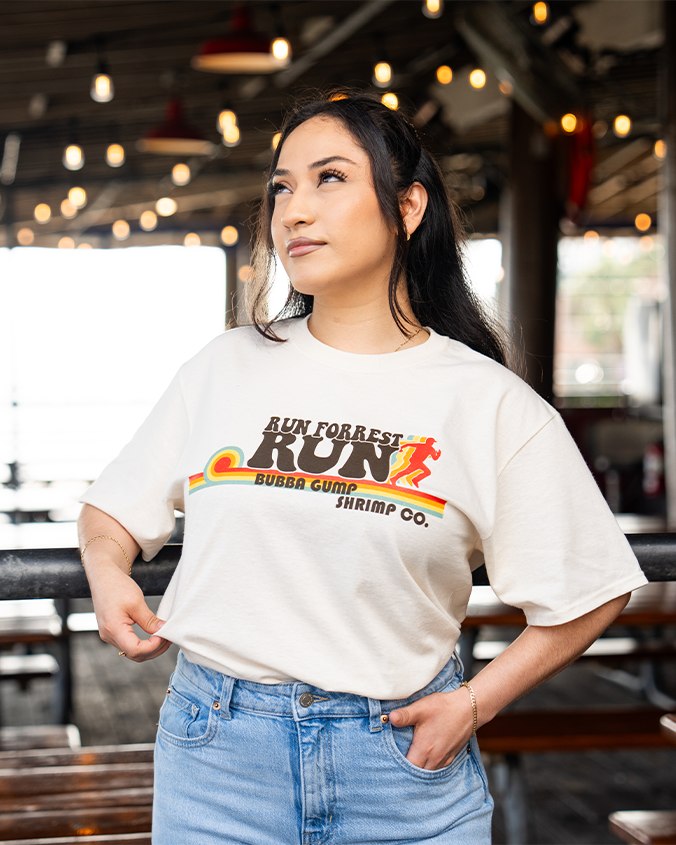 A dark-haired model with her hand in her pocket, in front of a restaurant setting wearing blue jeans tucked into an off-white t-shirt featuring the text 'RUN FORREST RUN' in bold letters above 'BUBBA GUMP SHRIMP CO.' The design includes a stylized running figure and horizontal stripes in rainbow.