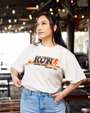 A dark-haired model with her hand in her pocket, in front of a restaurant setting wearing blue jeans tucked into an off-white t-shirt featuring the text 'RUN FORREST RUN' in bold letters above 'BUBBA GUMP SHRIMP CO.' The design includes a stylized running figure and horizontal stripes in rainbow.