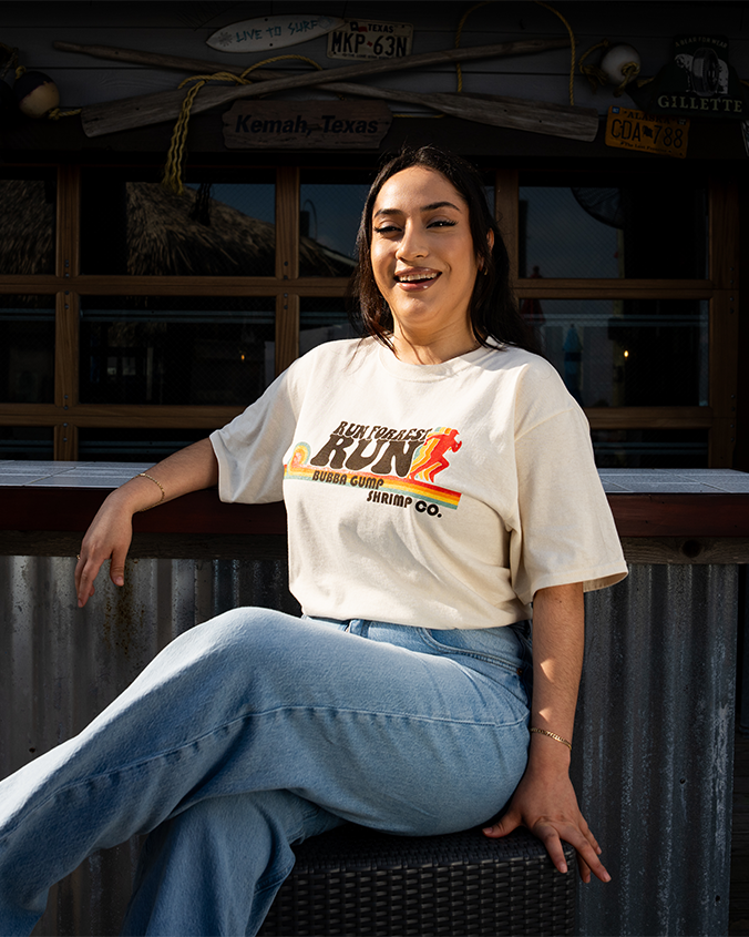 A dark-haired model sitting with her legs crossed on a stool in front of a set of rustically decorated wooden windows wearing blue jeans tucked into an off-white t-shirt featuring the text 'RUN FORREST RUN' in bold letters above 'BUBBA GUMP SHRIMP CO.' The design includes a stylized running figure and horizontal stripes in rainbow.