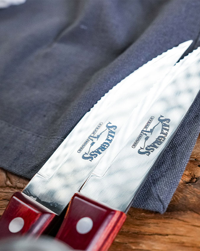 A close up of two steak knives blades showcasing engravement reading "SALTGRASS STEAKHOUSE", with wooden handles. The knives are placed against a grey towel on a wooden surface.