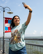 A dark-haired model taking a selfie on a pier in front of a blue and red banner on a pole, wearing blue jeans, gold jewelry and a blue t-shirt with a design featuring a selfie shot of multiple animals of the Savanna with cute funny faces against a sunny sky.