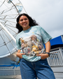 A person stands in front of a Ferris wheel, wearing a light blue shirt featuring a collage of various wild animals of the Savanna with cute funny faces.