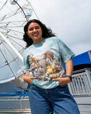 A dark-haired model standing in front of a Ferris wheel, wearing a blue t-shirt with a design featuring a selfie shot of multiple animals of the Savanna with cute funny faces against a sunny sky. She is also wearing blue jeans, a gold necklace, earrings, and bracelets.
