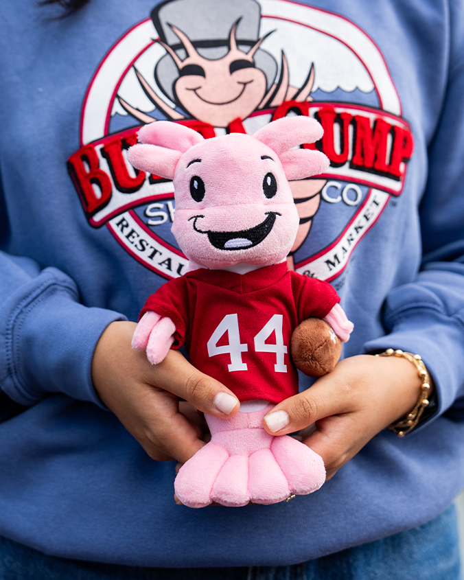 A close-up chest-level image of a model wearing blue jeans, gold bracelets, and a blue circular logo "Bubba Gump Shrimp Co." sweatshirt, holding up a pink plush smiling shrimp in a red sports jersey reading "44".