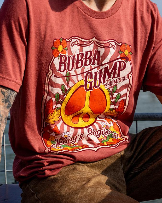 A close-up view of a sitting models' torso wearing brown pants and a crewneck t-shirt with the text "BUBBA GUMP Shrimp Co." and "Jenny's Singalong" in cursive with a large peace symbol and floral designs placed inside and outside of a white frame. The background is plain white.