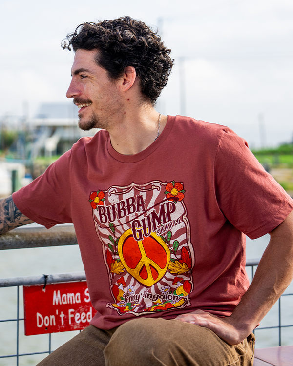 A young man sitting against a railing above a lake and sky, wearing a crewneck t-shirt with the text "BUBBA GUMP Shrimp Co." and "Jenny's Singalong" in cursive with a large peace symbol and floral designs placed in a white frame with stripes inside and flowers around the edges. 