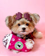 A small dog with a brown and black coat is sitting against a pink background. The dog is wearing a white outfit with black spots and pink trim, resembling a dress, and has a large pink bow on its head. A Starbucks Puppermint Plush is placed between her paws.