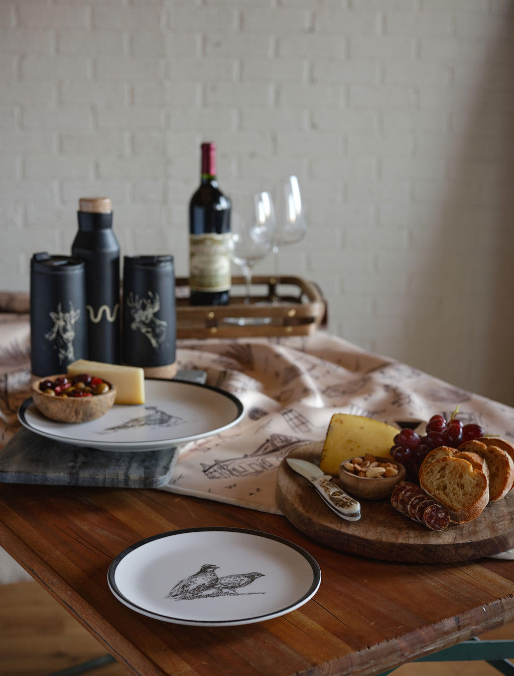 A rustic table setting featuring two plates with bird illustrations, a wooden charcuterie board, bowls and bottles with cork stoppers and deer illustrations, and a bottle of red wine, and two empty wine glasses on a wooden tray.